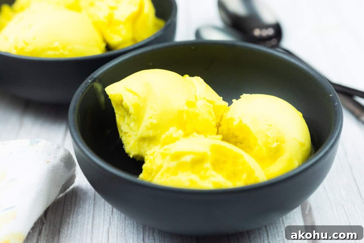 A black bowl containing three scoops of bright yellow ice cream sits on a light wooden surface, with another bowl and metal spoons in the background.