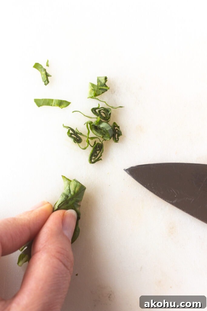 Close up of fresh basil leaves rolled together and being cut into small ribbons on a white cutting board.