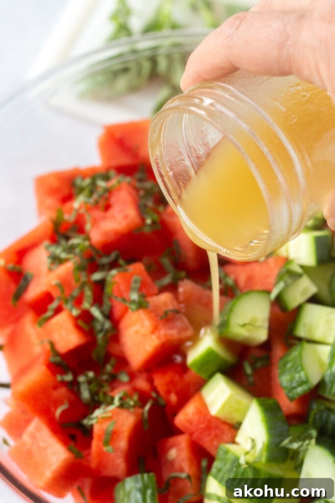Close up of golden salad dressing being poured over cut watermelon, chopped cucumber, and ribbons of fresh basil in a large bowl.