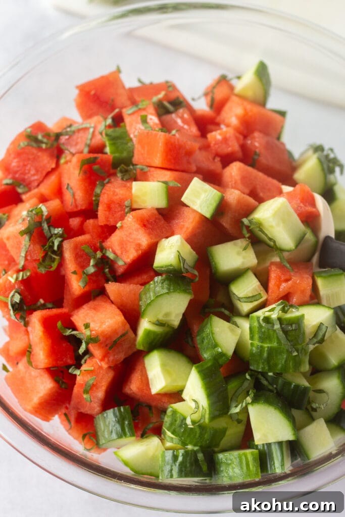 Close up of a rubber spatula stirring together the watermelon basil and cucumber salad in a large clear bowl.