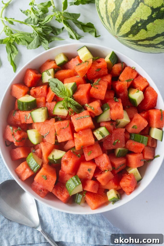 Top down shot of a large white bowl with a watermelon cucumber basil salad in it, surrounded by fresh basil, a watermelon, a blue cloth napkin, and a silver serving spoon.