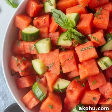 Close up of a bowl of watermelon salad with basil and cucumber in it. The bowl sits on a white background with fresh basil to the top left and a serving spoon in the bottom right of the frame.