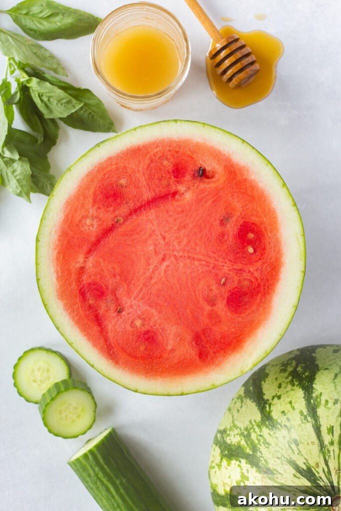 Top down shot of half a medium watermelon with fresh basil, sliced cucumber, and a light honey-colored dressing next to it, all on a white background.