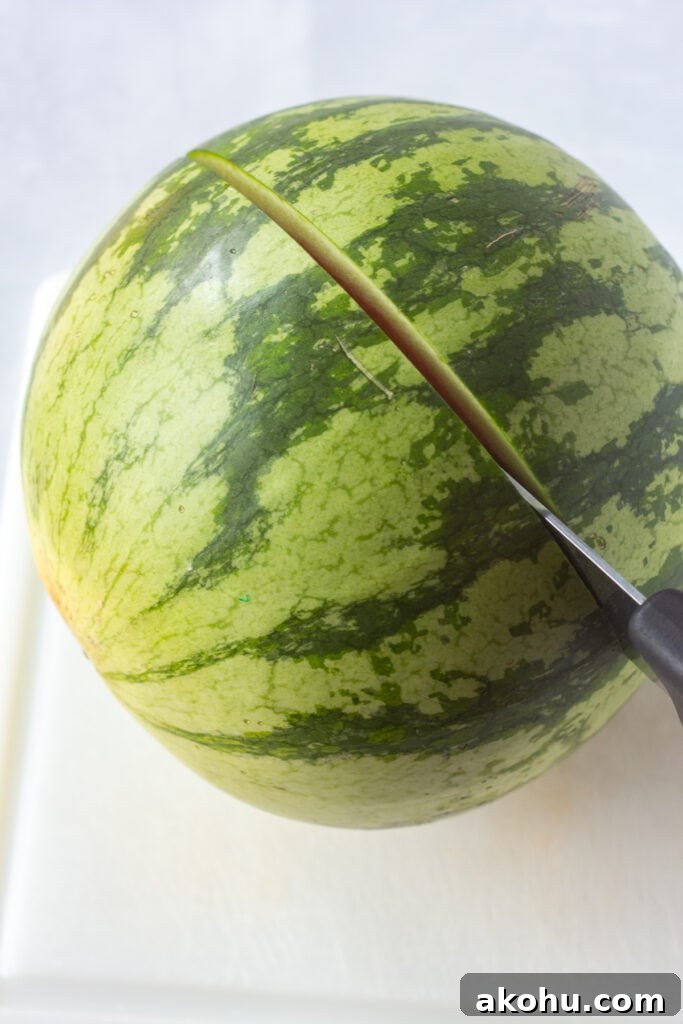 Close up of a medium-sized watermelon being cut in half with a large knife on a white cutting board.