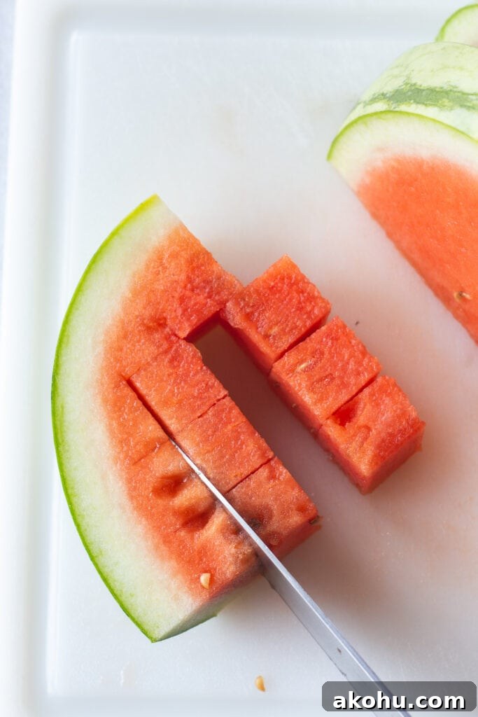 Close up of a quartered slice of a watermelon being cut into cubes by a large knife, all on a white cutting board.