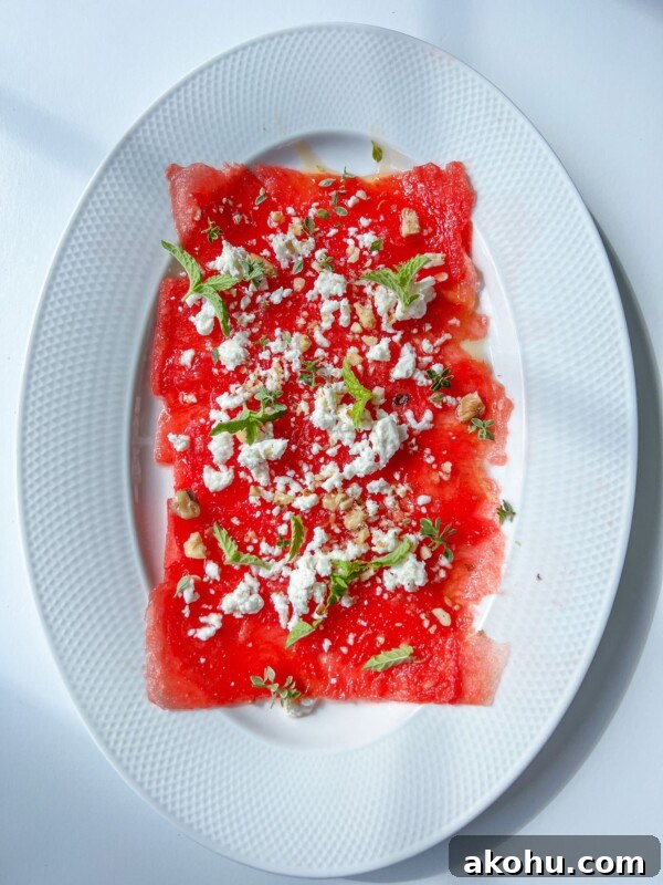 Watermelon Carpaccio: Thin slices of watermelon on an oval white plate, topped with crumbled white cheese, mint leaves, and a sprinkle of nuts or seeds, set against a white background.