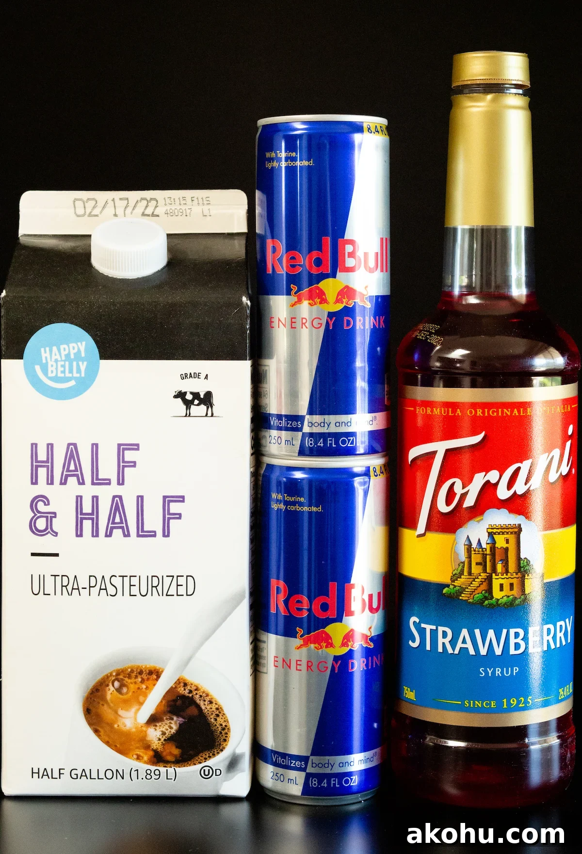 How to Make Sparkling Red Bull Italian Soda at Home 4 A carton of half and half, two cans of red bull, and a strawberry syrup bottle on a black background.