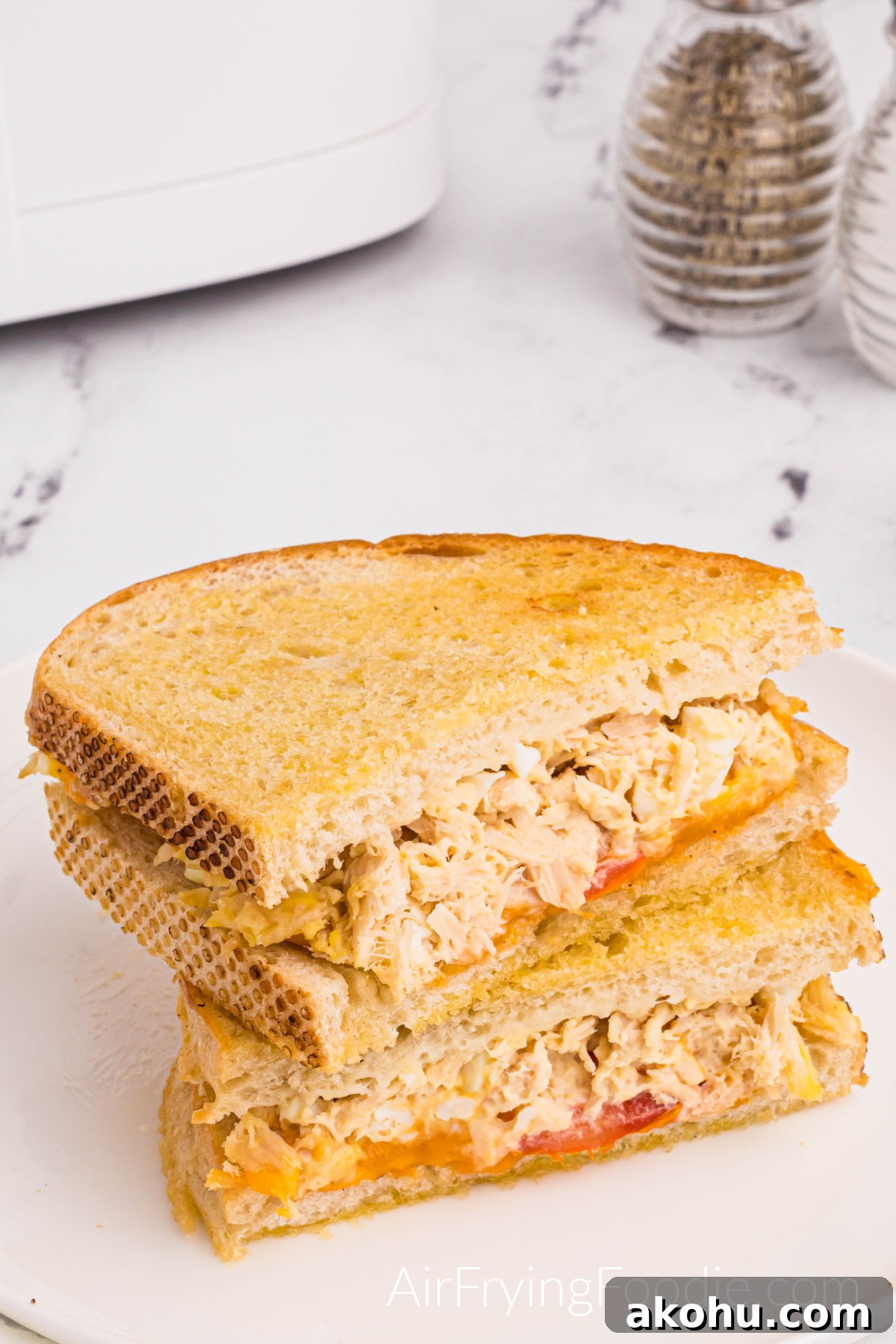 Tuna melt on golden Texas toast cooked in the air fryer with tomato and cheese cut in half and served on a round white plate. A small portion of the air fryer base is in the top left of the picture, and a sale and pepper shaker is in the top right. 