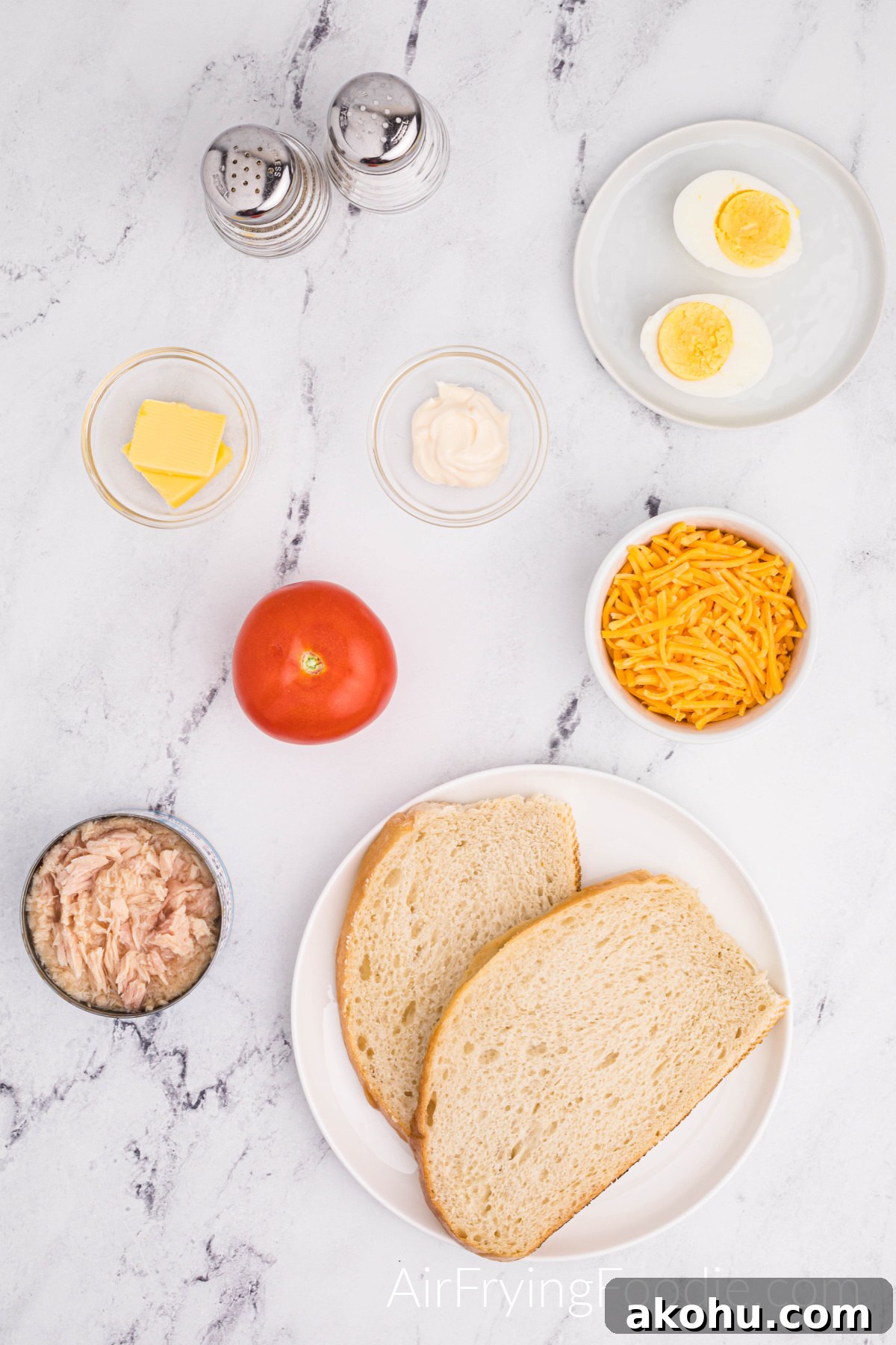 Crispy Air Fryer Tuna Melt Sandwich Recipe 3 Overhead shot of the ingredients used to make a tuna melt in the air fryer. Two round white plates; one with two slices of Texas toast and the second with one egg boiled and cut in half. Three small glass bowls contain tuna; the other has butter and mayonnaise—a small white bowl of shredded cheese, one whole tomato, and a salt and pepper shaker.