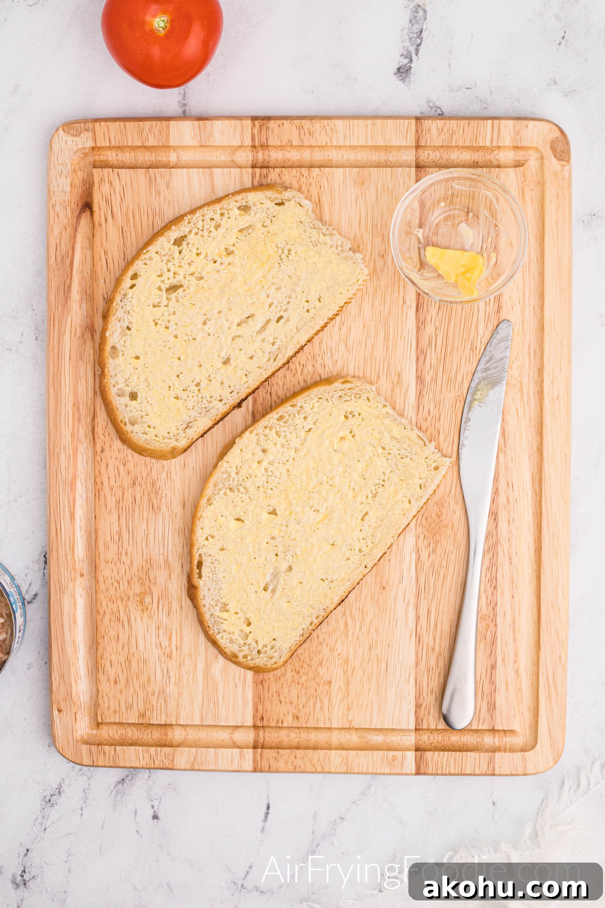Crispy Air Fryer Tuna Melt Sandwich Recipe 4 Two slices of Texas toast buttered on a cutting board with a small glass bowl of butter and a butter knife. There is a tomato to the top left of the cutting board.