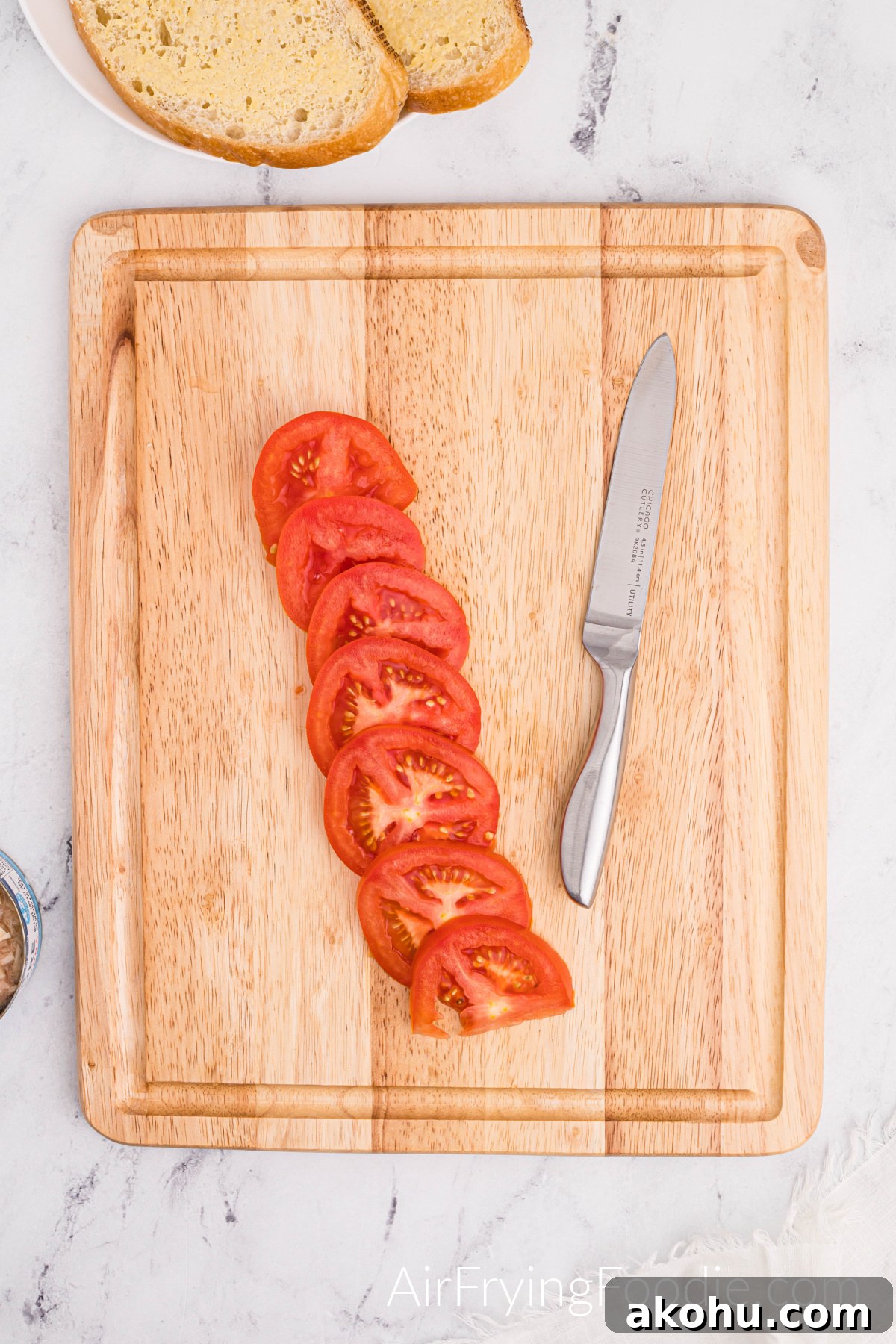 Crispy Air Fryer Tuna Melt Sandwich Recipe 5 A tomato is sliced and lined on a cutting board with a butter knife on the right of the cutting board and two slices of Texas toast on a round white plate above the cutting board.