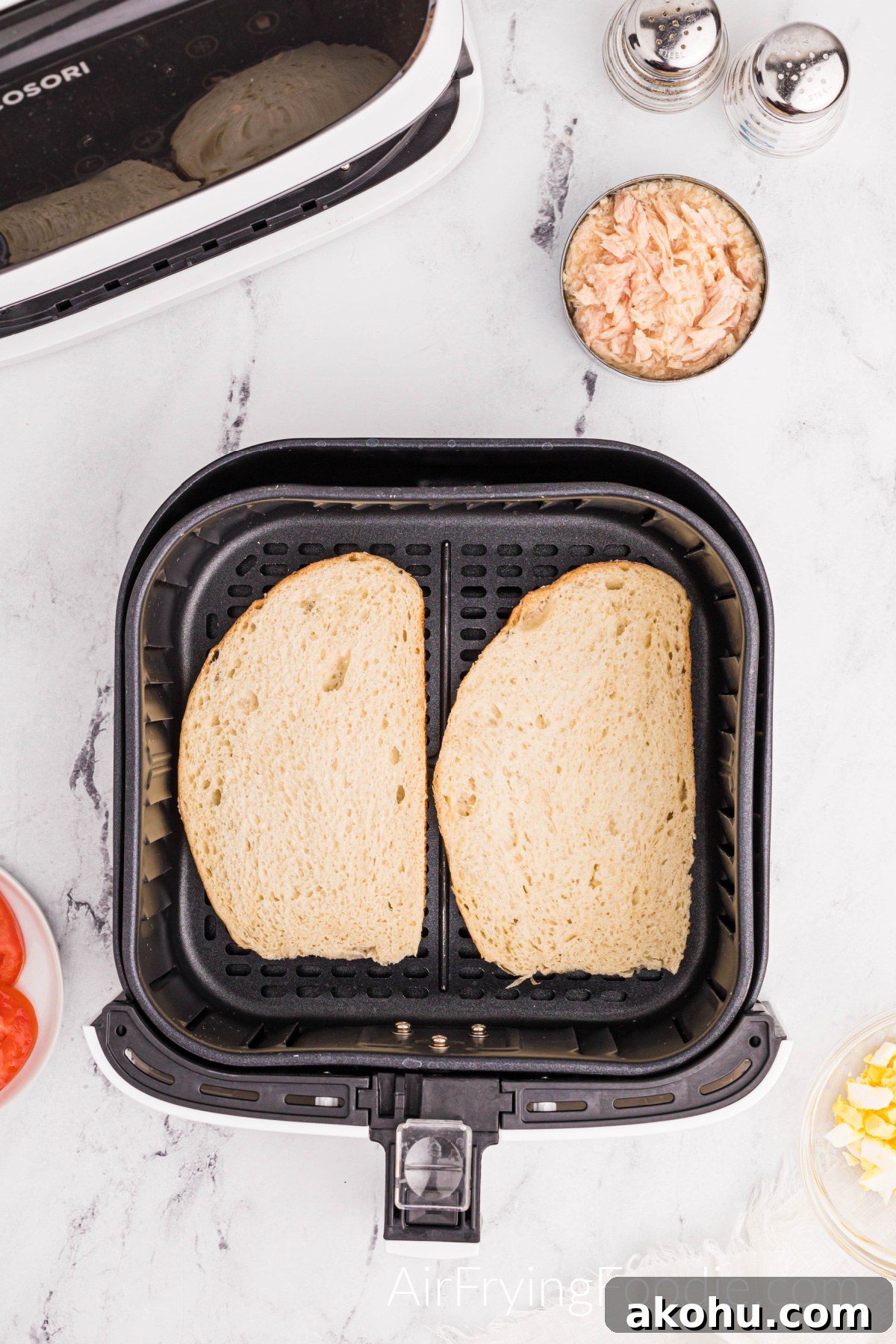 Crispy Air Fryer Tuna Melt Sandwich Recipe 6 Two slices of Texas toast in the air fryer with a salt and pepper shaker and a glass bowl of tuna to the top right of the air fryer basket. The air fryer base is in the top left of the image. A small portion of the tomato is shown to the left of the air fryer basket, and a small amount of a glass bowl with eggs is to the right of the air fryer basket.