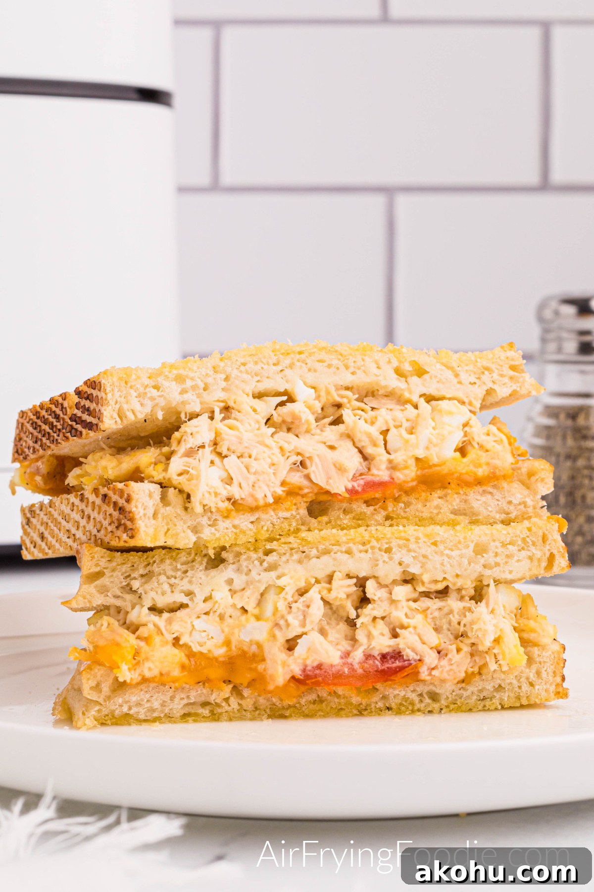Tuna melt sandwich on golden Texas toast cooked in the air fryer with tomato and cheese cut in half and served on a round white plate. There is a small portion of a pepper shaker to the right of the sandwich and a small part of the air fryer base to the left of the picture. 