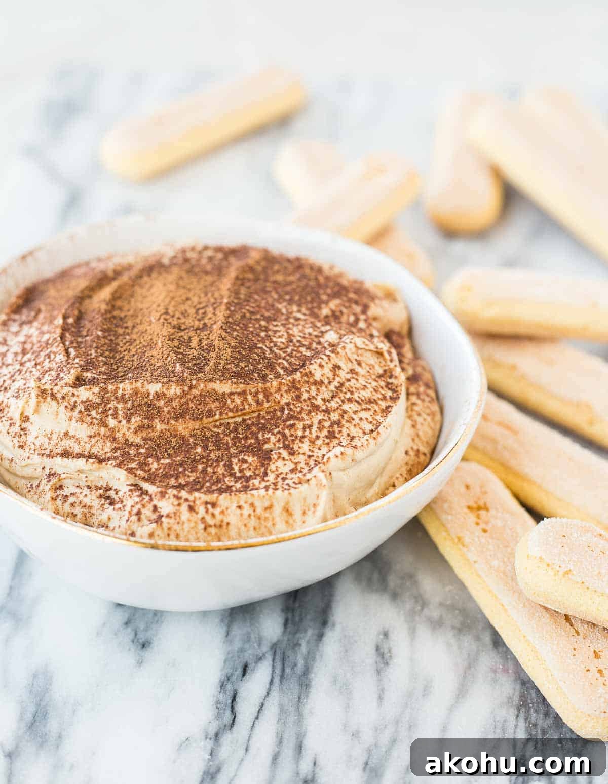 10-Minute Tiramisu Dip: Creamy Espresso Mascarpone Recipe 2 Closeup of tiramisu dip in a round white bowl, garnished with cocoa powder, and surrounded by ladyfingers, on a white marble surface.