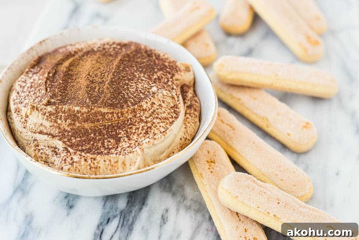10-Minute Tiramisu Dip: Creamy Espresso Mascarpone Recipe 4 Tiramisu dip in small white bowl, garnished with a dusting of cocoa powder, with lady fingers arranged around it.