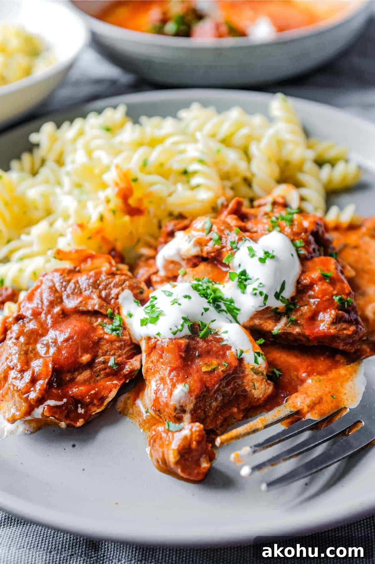 A plate of Hungarian Pörkölt Beef Stew and pasta fusilli drizzled with sour cream and garnished with parsley.