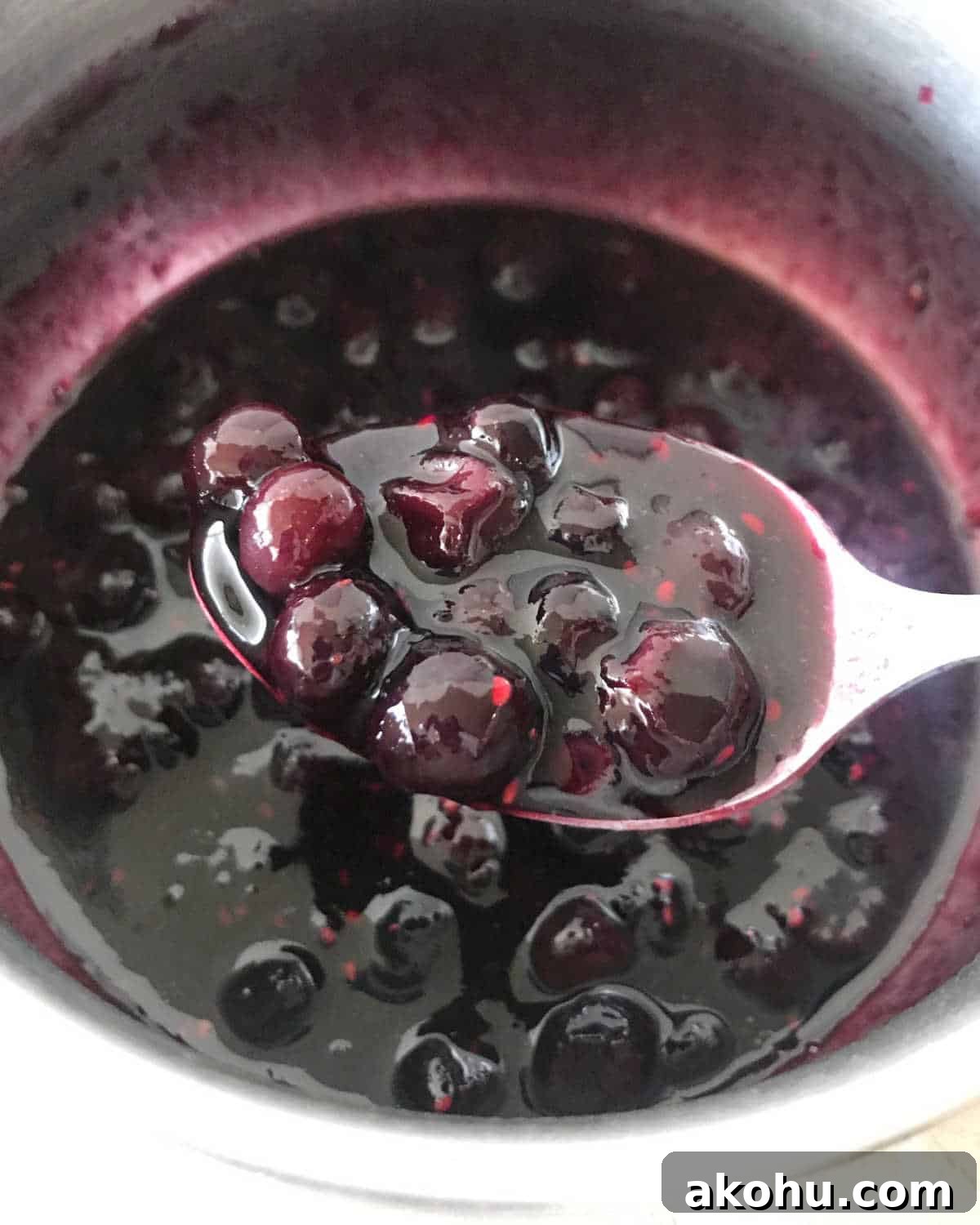 Blueberry Sauce Recipe for Pancakes, Yogurt & Desserts 5 Silver spoon with blueberry sauce over metal saucepan where it's being cooked.
