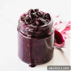 Blueberry Sauce Recipe for Pancakes, Yogurt & Desserts 9 Glass jar with blueberry sauce, a spoon, white background.