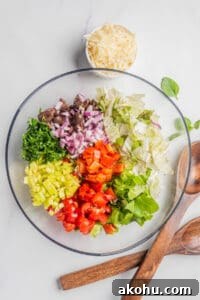 An image of Romaine lettuce, endive, red onion, cherry tomatoes, cucumber, pepperoncini, roasted red peppers, olives and parsley in a large mixing bowl. 
