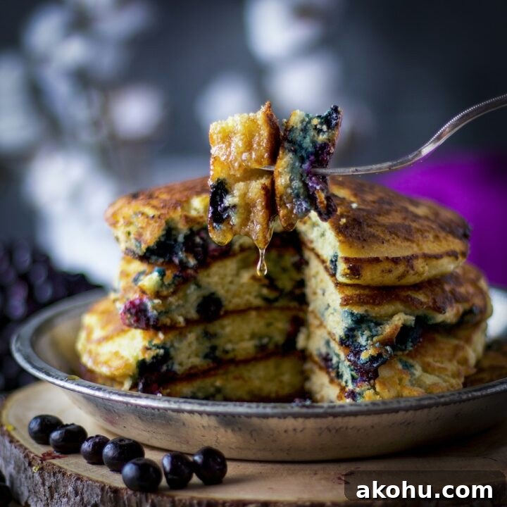 A fork with a bite of fluffy blueberry pancakes on it, syrup dripping from the pancakes, and a stack of blueberry pancakes in the background.