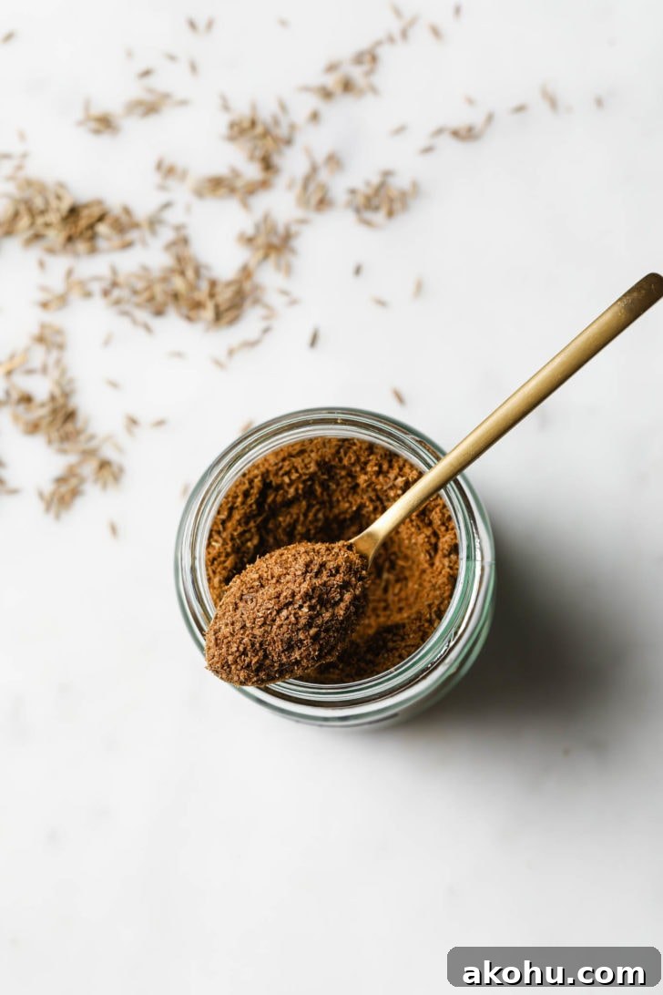 A gold spoon with cumin powder resting on top of a jar of cumin powder.