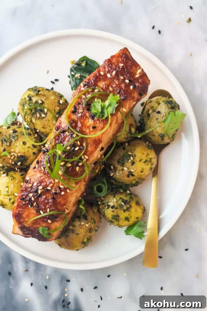 Miso glazed salmon on top of herby potatoes on a white plate, with sesame seeds, coriander and spring onion on top.