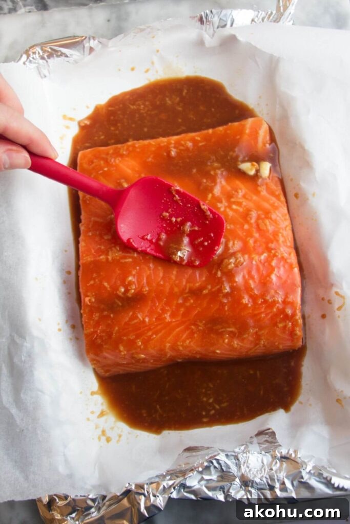 Small red spatula spooning maple miso glaze over a salmon fillet in a baking paper lined tray.