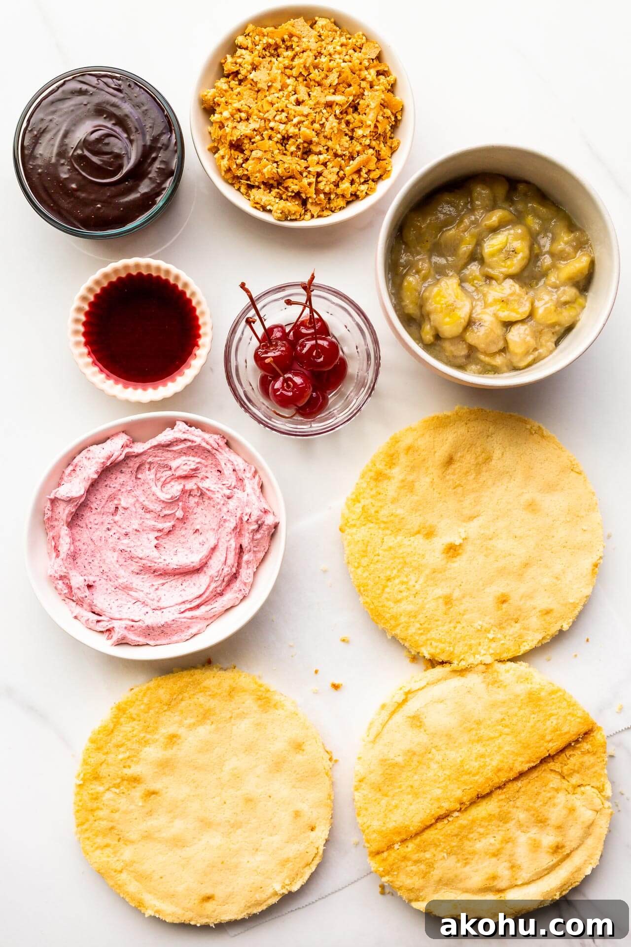 The components of a banana split cake, ready to be stacked and assembled into a layer cake, including three layers of vanilla cake, cherry frosting, maraschino cherries, cherry juice, banana compote, peanut crunch, and chocolate fudge sauce.