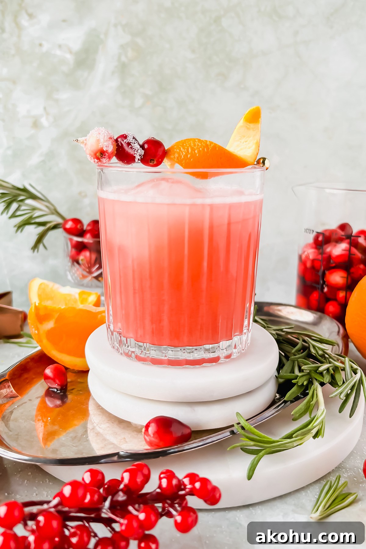 cranberry whiskey sour garnished with orange peel and cranberries on white coaster on silver tray surrounded by additional cocktail garnishes.
