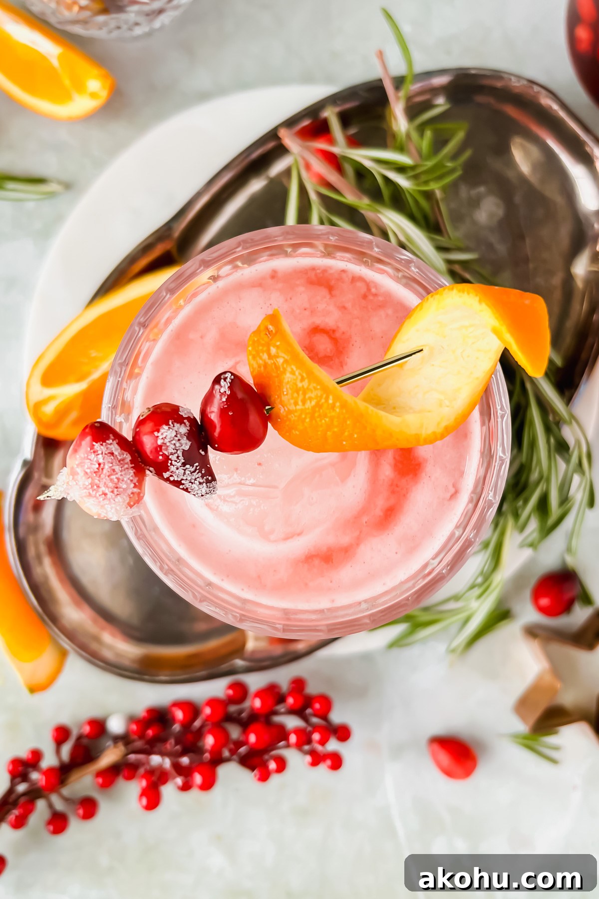 overhead shot of cranberry whiskey sour garnished with orange peel and cranberries on white coaster on silver tray surrounded by additional cocktail garnishes.