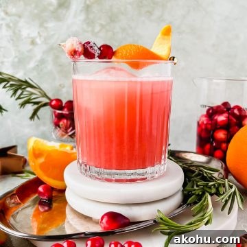 cranberry whiskey sour garnished with orange peel and cranberries on white coaster on silver tray surrounded by additional cocktail garnishes.