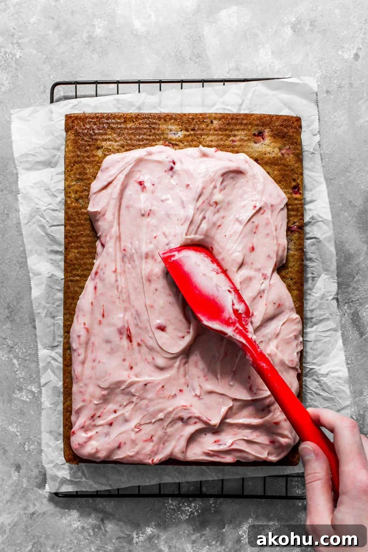 strawberry sheet cake on parchment lined wire rack with frosting being spread on top