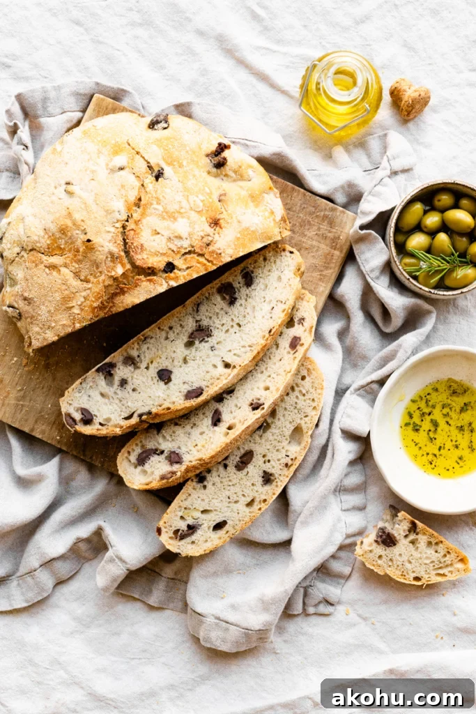 No-Knead Olive and Herb Artisan Bread Recipe 2 Overhead view of a loaf of olive bread on a wood cutting board with three slices cut. The board is on a linen covered table next to bowls of olives and olive oil.
