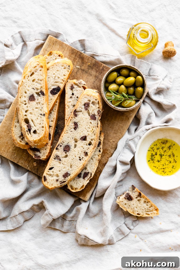 No-Knead Olive and Herb Artisan Bread Recipe 12 Slices of olive bread stacked on a wood cutting boards next to a bowl of olives on a linen covered table with a bowl of olive oil.