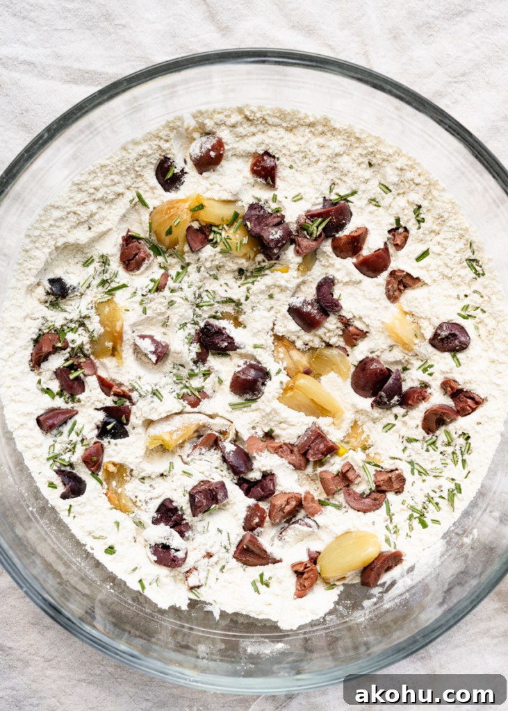 No-Knead Olive and Herb Artisan Bread Recipe 4 Overhead view of a bowl of flour with chopped kalamata olives, chunks of roasted garlic, and chopped rosemary.