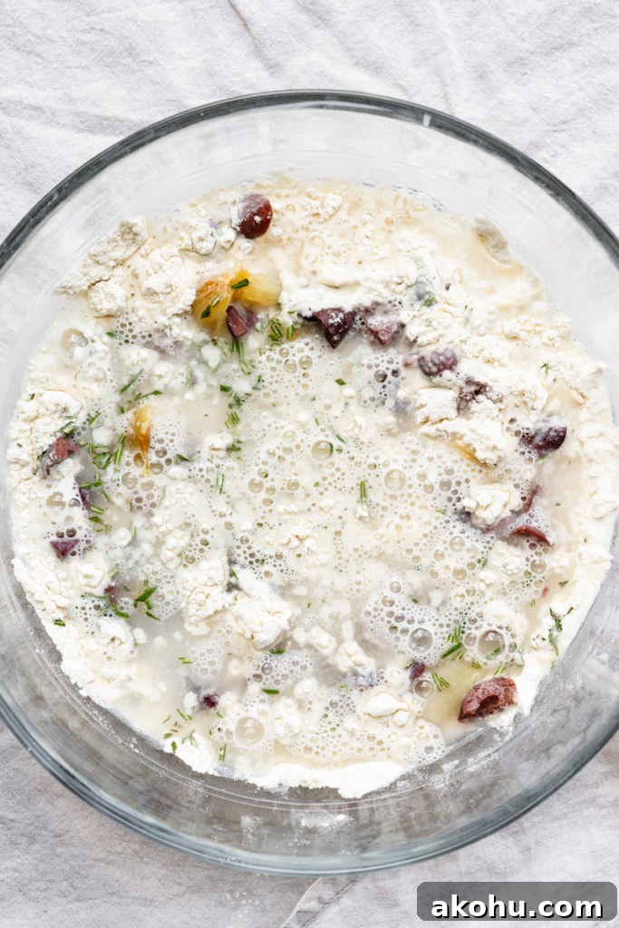 No-Knead Olive and Herb Artisan Bread Recipe 5 Overhead view of a bowl of flour with chopped kalamata olives, chunks of roasted garlic, chopped rosemary, and water.