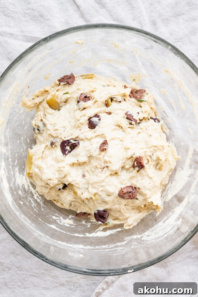 No-Knead Olive and Herb Artisan Bread Recipe 6 Overhead view of a bowl of bread dough with chopped kalamata olives, chunks of roasted garlic, chopped rosemary, and water.