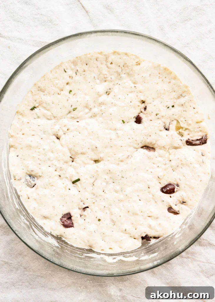 No-Knead Olive and Herb Artisan Bread Recipe 7 Overhead view of a bowl of bread dough with chopped kalamata olives, chunks of roasted garlic, chopped rosemary, and water.