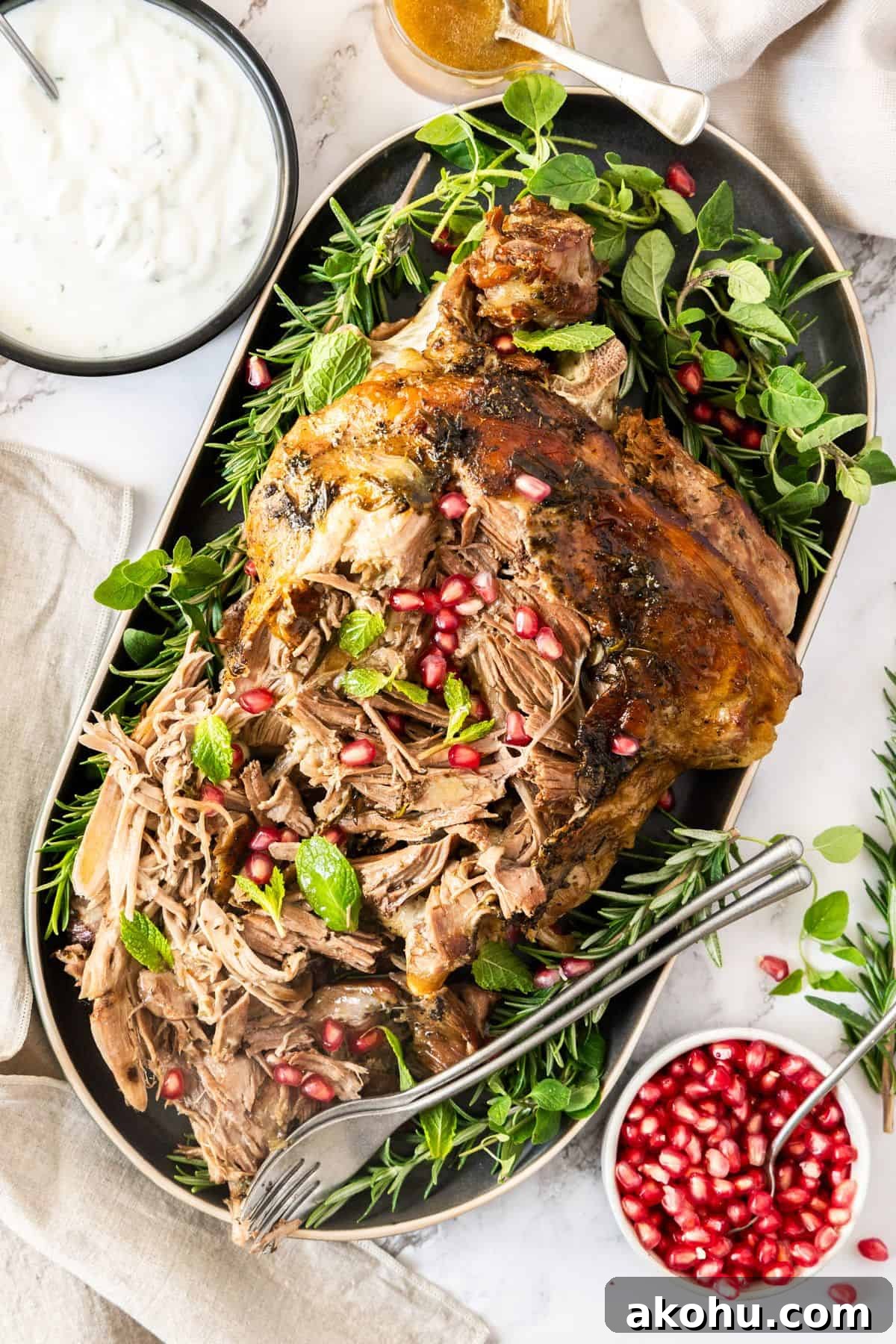 Easter Dinner Mains: Elegant Roast and Ham Recipes 17 Slow Roasted Leg of Lamb, surrounded by fresh herbs and pomegranate, on a grey oval platter, with two forks on the edge.