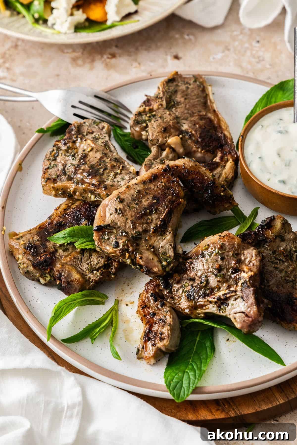 Easter Dinner Mains: Elegant Roast and Ham Recipes 19 Round white plate with cooked Marinated Lamb Loin Chops, with a small bowl of yoghurt sauce on the side, and white cloth.