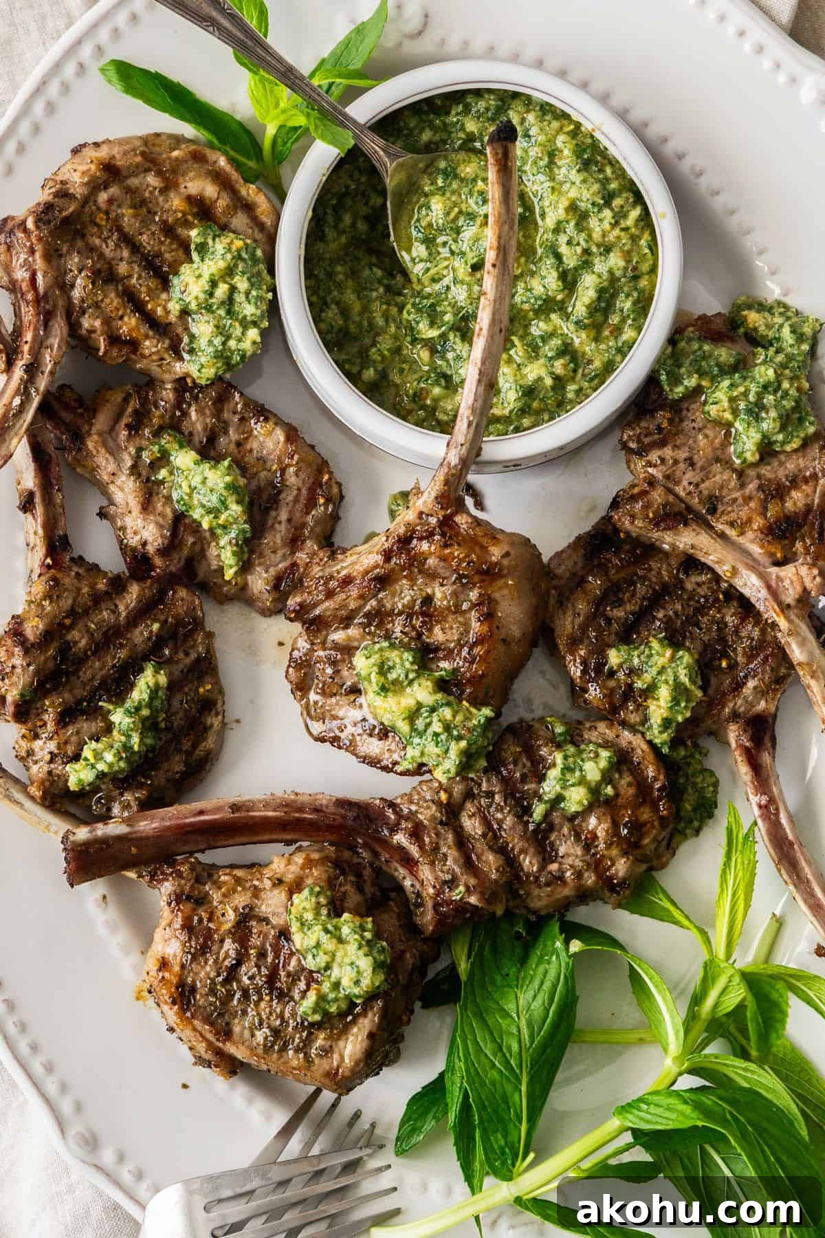 Easter Dinner Mains: Elegant Roast and Ham Recipes 9 Oval white plate with Grilled Lamb Chops, some fresh mint leaves, two forks and a small dish of Mint Pesto.