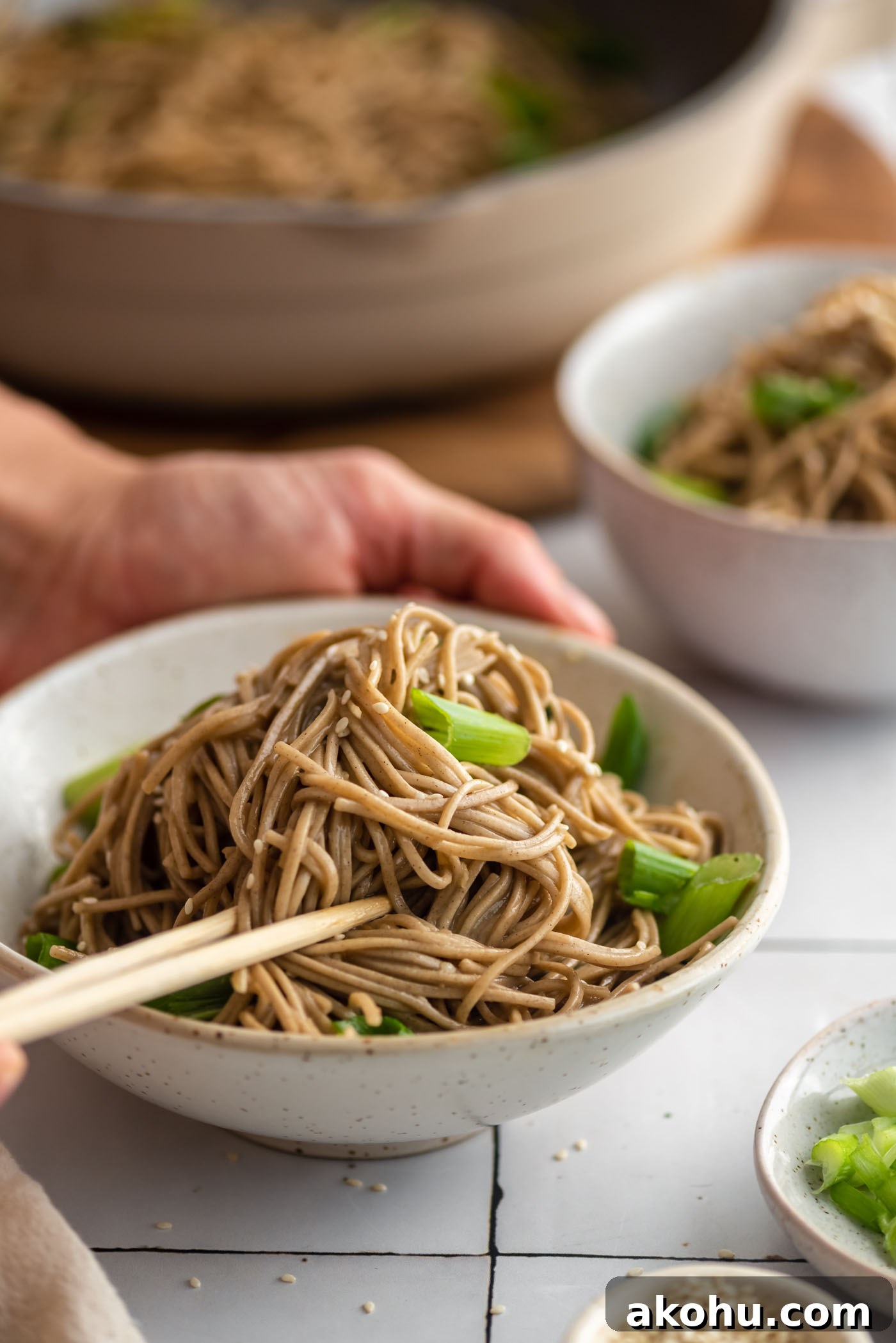 Two hands using chopsticks in a bowl of cold sesame noodle salad with green onion and sesame seeds.