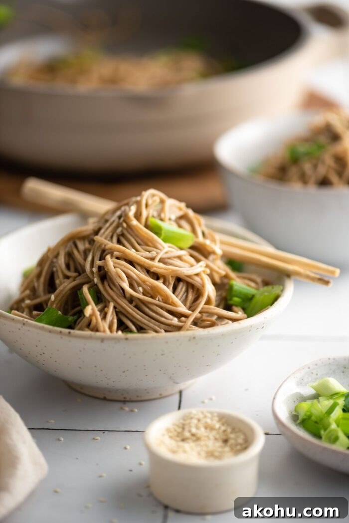 Bowl of sesame buckwheat soba noodle salad with green onions, topped with sesame seeds. A pair of chopsticks rests on the side of the bowl.