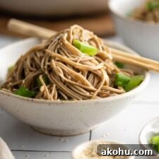 Bowl of sesame buckwheat soba noodle salad with green onions, topped with sesame seeds. A pair of chopsticks rests on the side of the bowl.