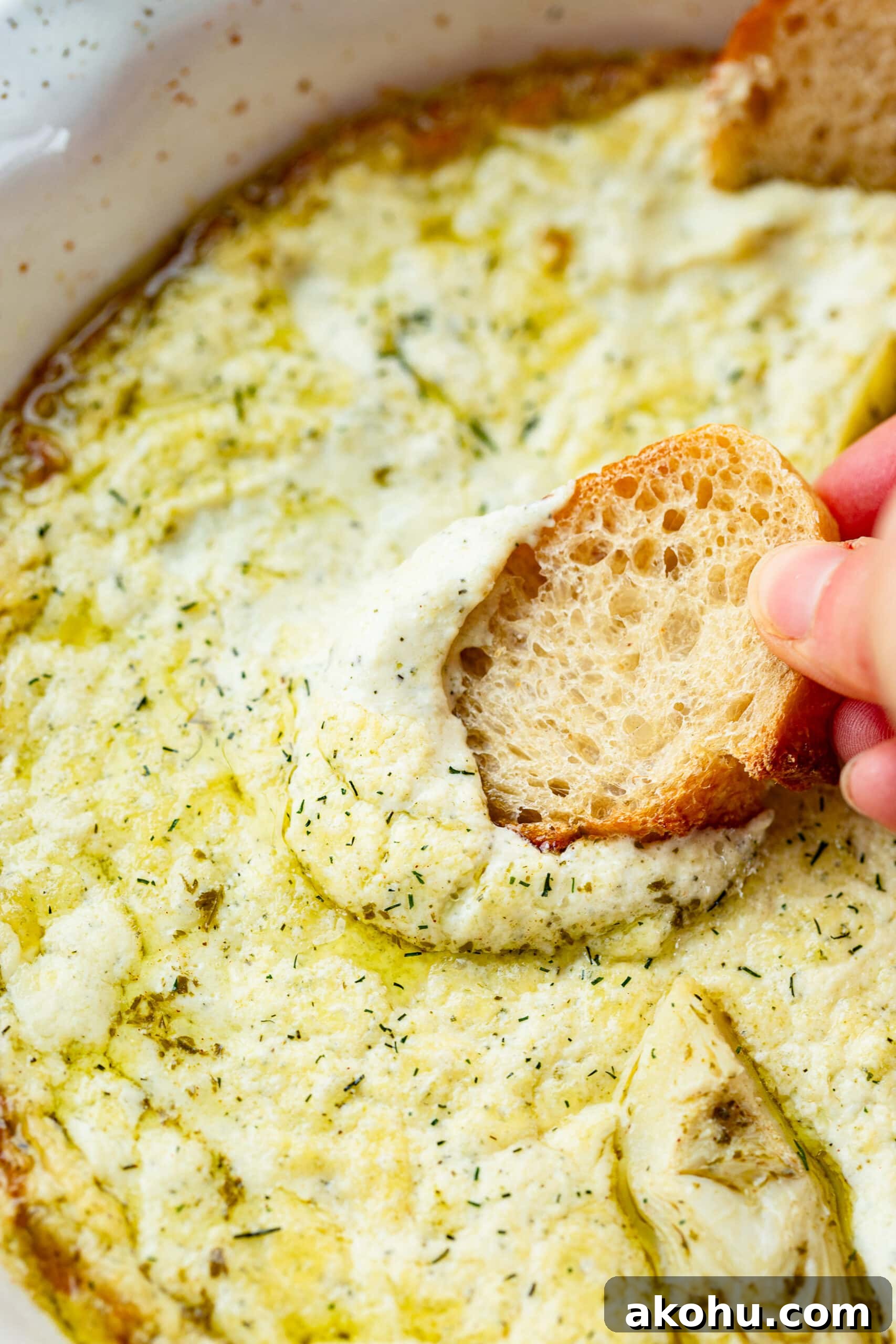 a hand dipping a crostini in a large dish filled with creamy artichoke dip.