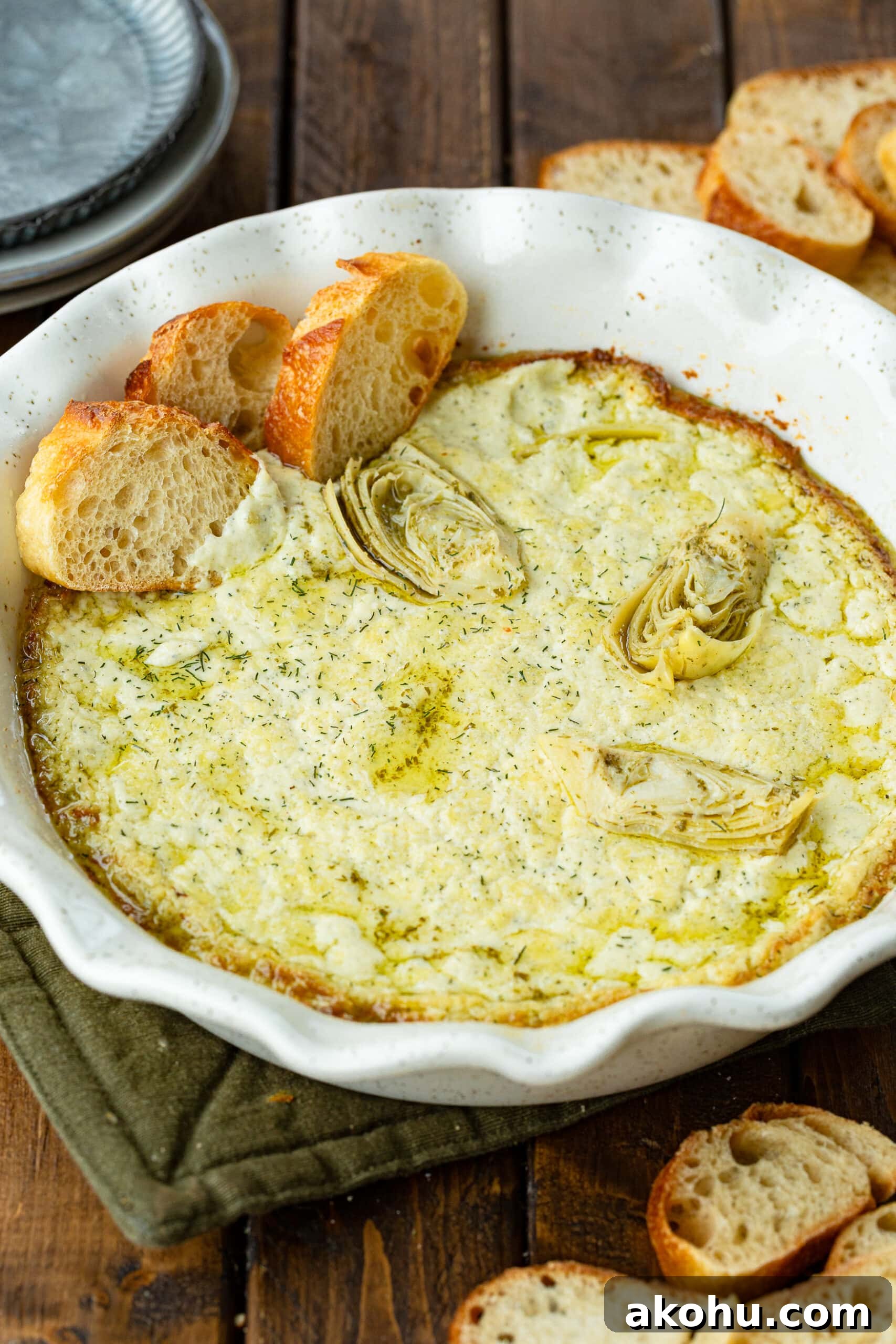 a circular white ceramic baking dish full of golden creamy artichoke dip with three slices of baguette stuck in the dip along the side of the pan.