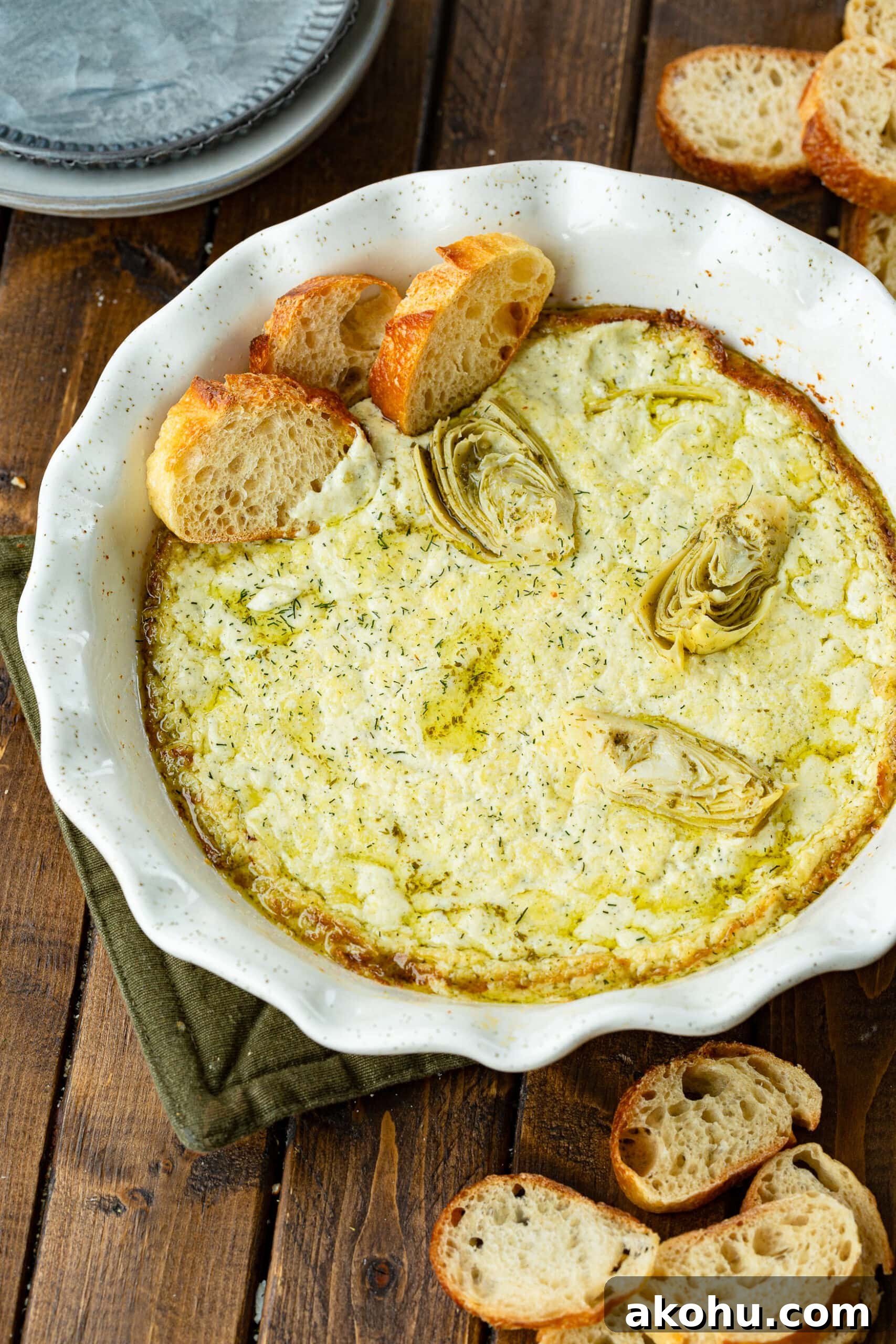 a round white baking dish full of creamy artichoke dip that is baked golden around the edges and has three slices of baguette sticking in it on the side of the dish.