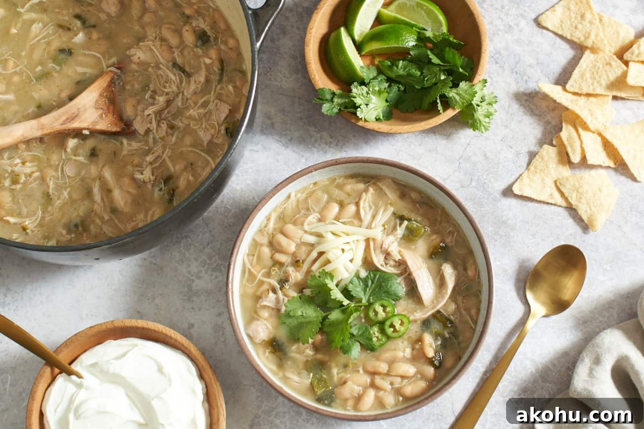 A bowl of white chicken chili topped with cheese, peppers and cilantro, surrounded by a pot of chili, bowls of sour cream, cilantro, and limes, and some tortilla chips.