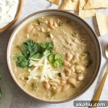 A bowl of white chicken chili with poblano peppers topped with cheese, chilis and cilantro, a bowl of sour cream is on the left, some tortilla chips and a gold spoon are on the right.