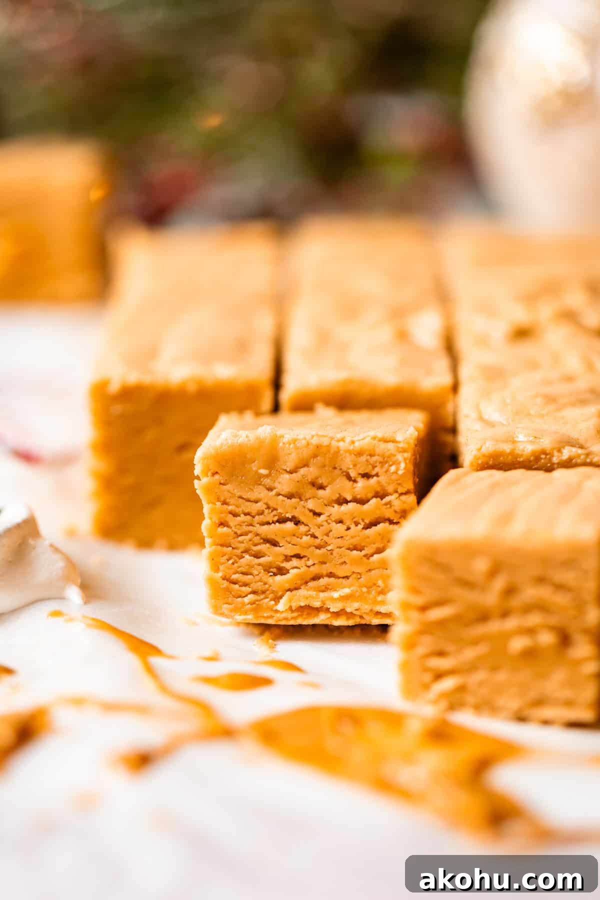 Close-up of several pieces of creamy, peanut butter fudge arranged on parchment paper, with smooth, textured surfaces and a blurred festive background.